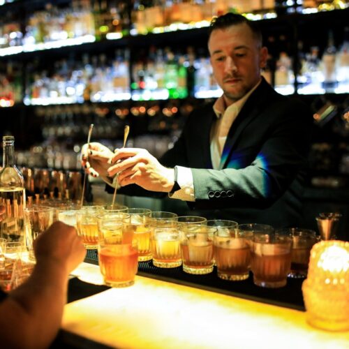A bartender pouring rows of cocktails at a backlit bar.