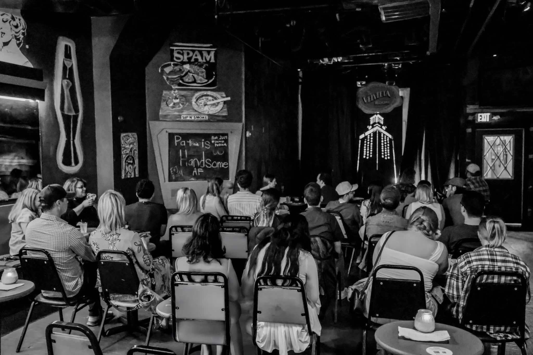 An audience seated at small tables watching a comedian on a club stage, in black and white.