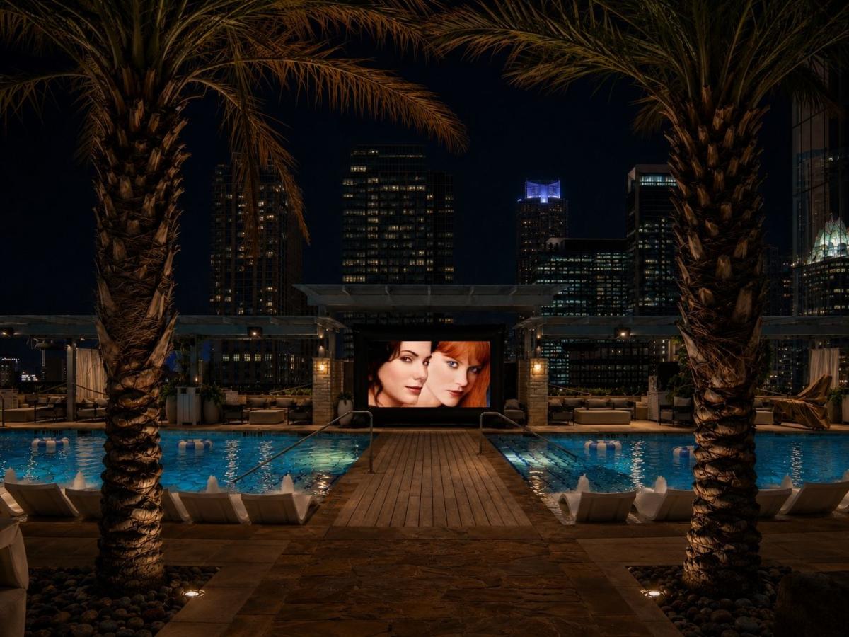 A movie playing on an outdoor screen at the end of a rooftop pool, framed by palm trees and downtown towers at night.