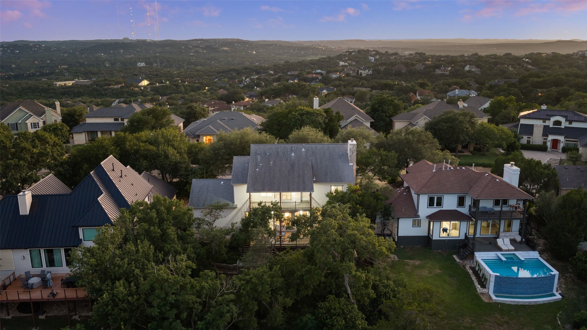 An aerial view at dusk over Northwest Hills rooftops and live oaks, with Hill Country ridges in the distance.