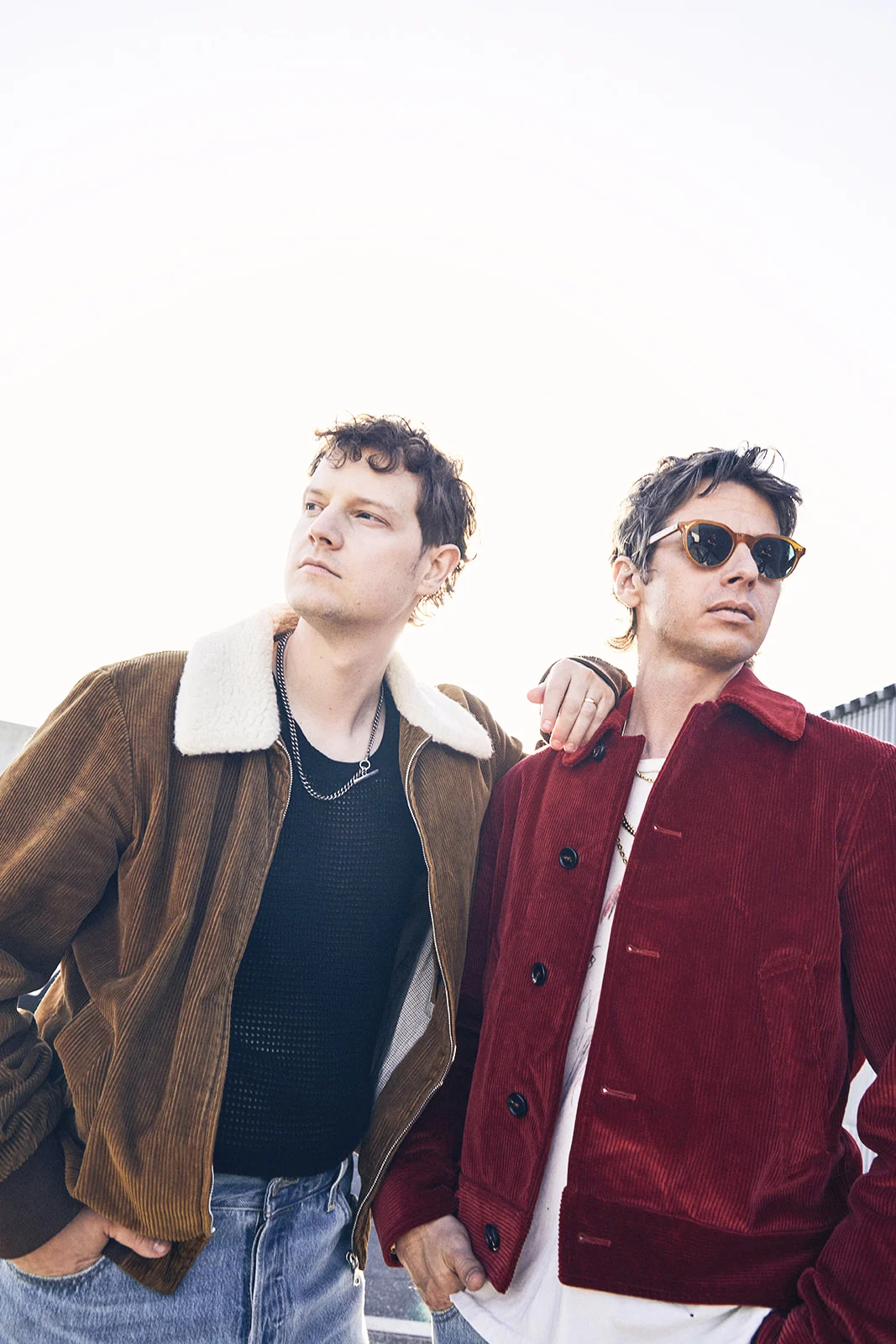 Two members of Foster the People stand outdoors in shearling and corduroy jackets against a pale sky.