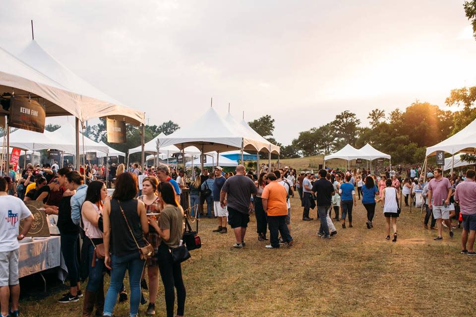 A crowd walks between white festival tents on a grassy field in the late afternoon.
