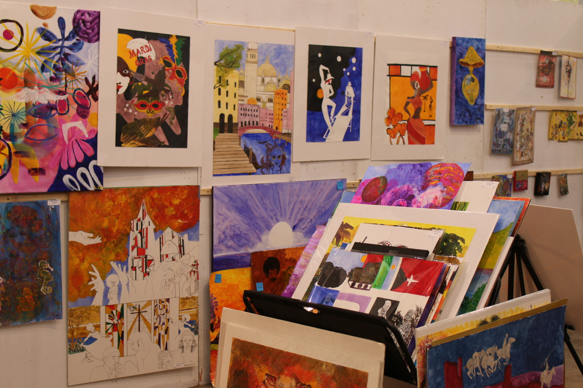 Colorful paintings hang on white gallery walls above a bin of framed prints.