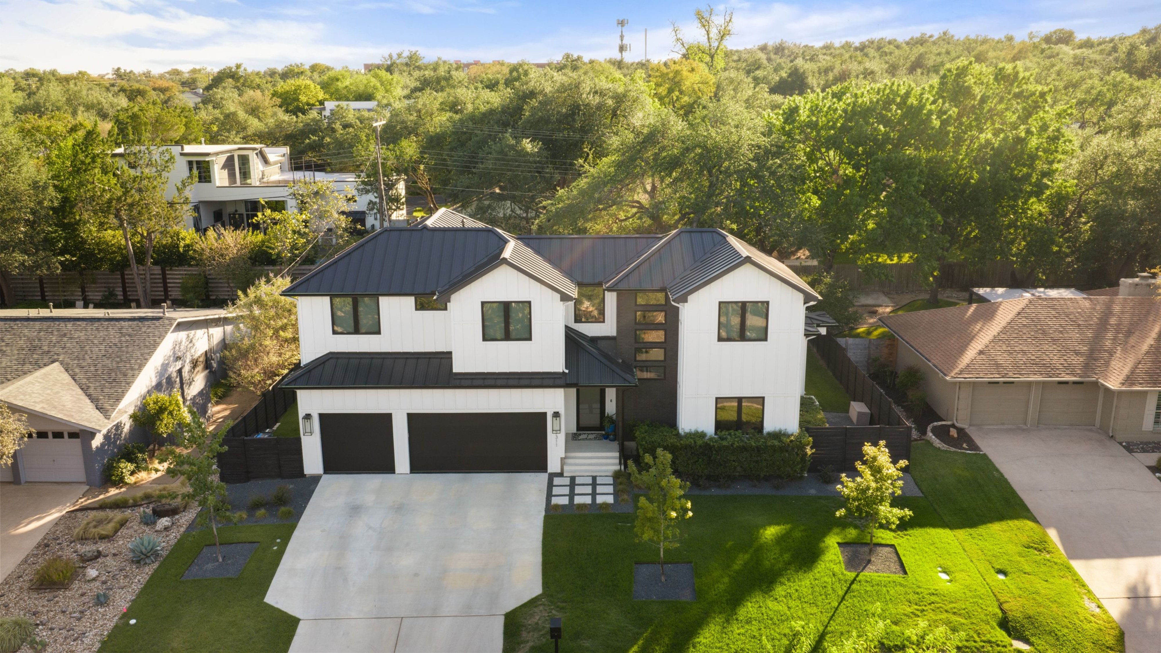 7311 Running Rope, a two-story white home with a dark metal roof and two garage doors, set among live oaks in Northwest Hills.