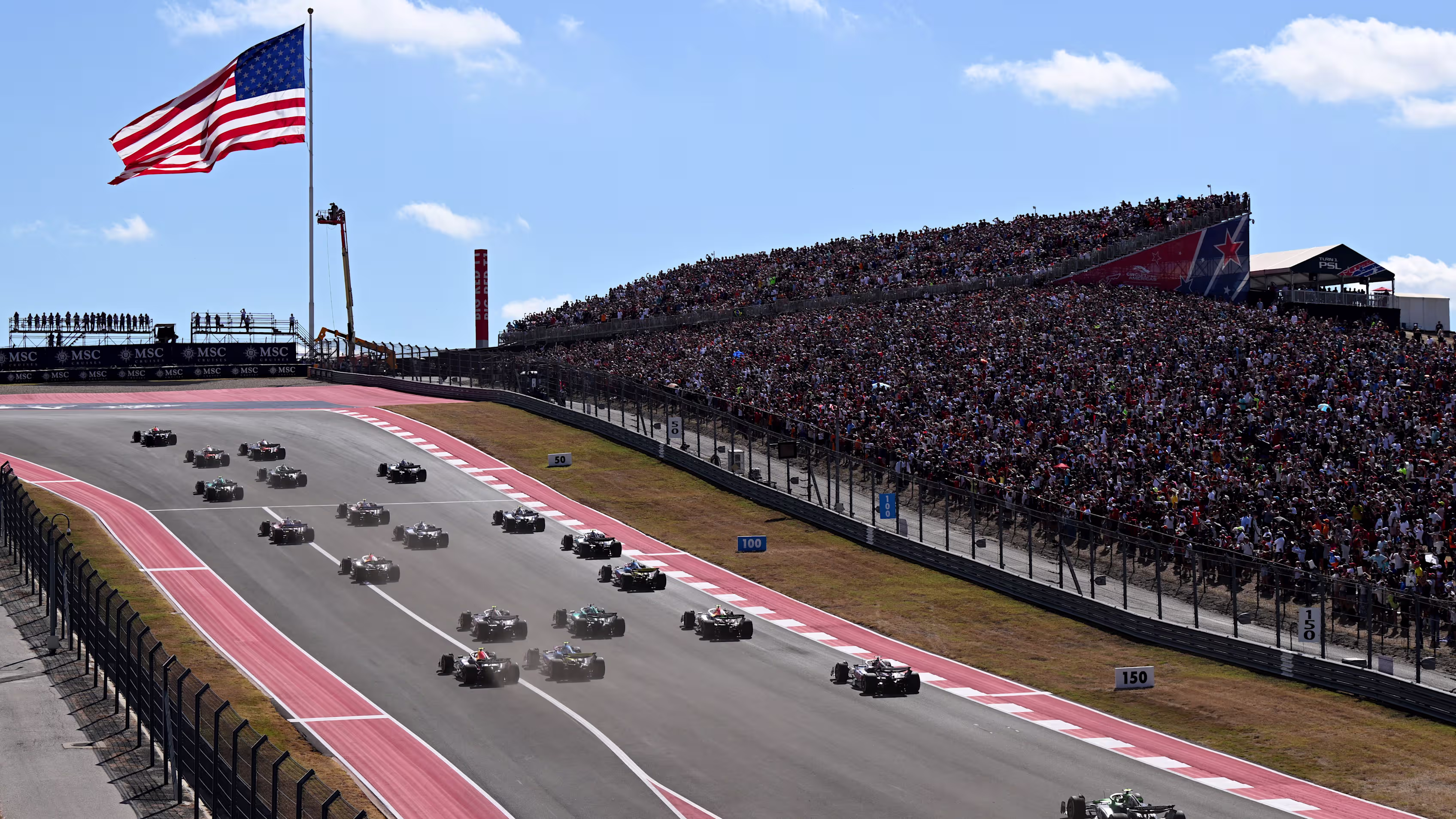 Formula 1 cars race down the front straight at Circuit of The Americas below packed grandstands and a large American flag.