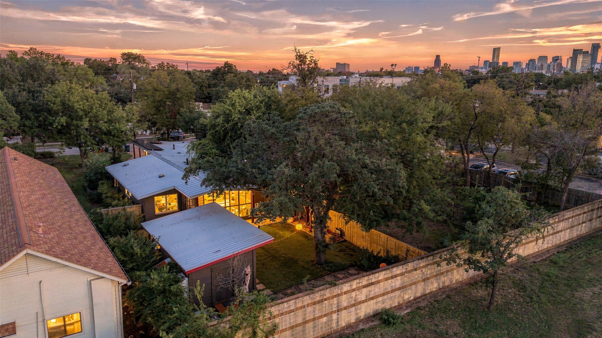An aerial view at sunset of 3504 West Avenue, a low home under mature trees, with the downtown Austin skyline beyond.