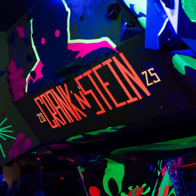 A neon Crank’N’Stein sign glows under black light on a climbing wall covered in fluorescent paint.